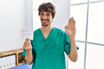 Poster - Young hispanic doctor man holding covid record card waiving saying hello happy and smiling, friendly welcome gesture