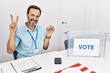 © Krakenimages.com - Middle age man with beard sitting by ballot holding i vote badge smiling looking to the camera showing fingers doing victory sign. number two.