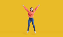 Happy Ecstatic Pretty Young Woman In Casual Wear Having Fun In Studio. Carefree Teenage Girl In Comfortable Orange Sweatshirt And Blue Jeans Jumping High For Joy Isolated On Vibrant Yellow Background