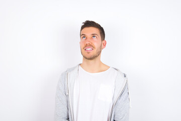 Wall Mural - Portrait of mysterious young caucasian man wearing casual clothes over white background looking up with enigmatic smile. Advertisement concept.