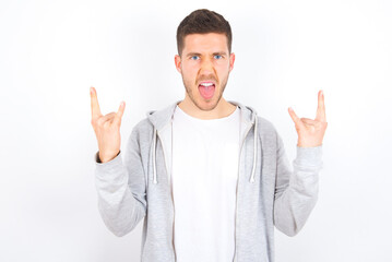 Born to rock this world. Joyful young caucasian man wearing casual clothes over white background screaming out loud and showing with raised arms horns or rock gesture.