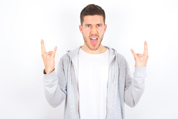 Wall Mural - Born to rock this world. Joyful young caucasian man wearing casual clothes over white background screaming out loud and showing with raised arms horns or rock gesture.