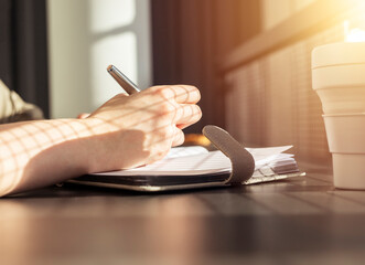 Poster - Female hands closeup taking notes in notebook. Woman sitting at table with collapsible silicon mug and writing agenda or thoughts in office or cafe with sunlight. High quality photo