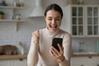 © fizkes - Happy young woman look at phone screen clenched fist, scream with joy, rejoicing success, get great unbelievable news in message or sms sit in kitchen at home. Moment of win, luck, bet victory concept