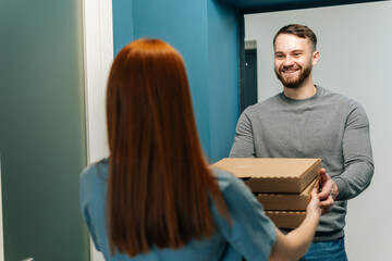 Cheerful bearded courier male delivering boxes with hot pizza to unrecognizable female customer on doorway at home. Back view of young woman client receiving boxes with food from delivery man.