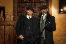 Two Men Of Different Ages, English Retro Gangster Of The 1920s Dressed In A Coat, Suit And Flat Cap In Peaky Blinders Style.