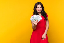 Smiling Business Woman Wears Red Dress Showing Fan Of Cash Money In Euro Banknotes In Yellow Background In Studio. Rich Girl Looks Aside. Business, Finance, Saving, Banking