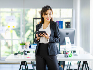 Asian young female professional successful businesswoman secretary employee staff in formal suit pants standing smiling holding touchscreen tablet computer checking email in company office workplace