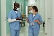 © pressmaster - Two young intercultural doctors or surgeons in protective masks having discussion of troubles while walking along hospital corridor