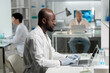© pressmaster - Side view of young African American researcher in lab coat and surgical gloves typing on laptop keyboard while sitting by desk