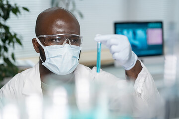 Aufkleber - Young black man in lab coat, protective mask and gloves looking at flask containing blue liquid during work in laboratory