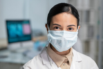 Aufkleber - Young pretty female clinician in whitecoat and protective mask on face looking at camera while standing in medical office