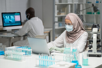 Aufkleber - Muslim female scientist in hijab and protective workwear typing on laptop keyboard while sitting by workplace against coworker