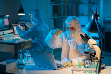 Aufkleber - Young contemporary chemist in biohazard suit studying liquid substance while sitting by desk in front of laptop in laboratory
