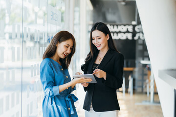 Smiling businesswoman holding digital tablet and talking with partner while standing in modern office interior, team of professional employees discussing ideas of project after working on touch pad