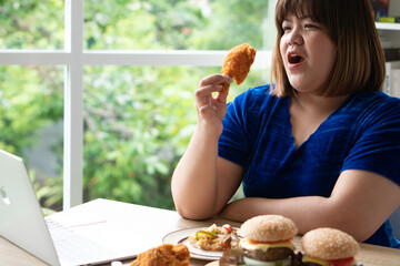 Hungry overweight woman holding Fried Chicken, hamburger on a wooden plate and Pizza on table, During work from home, gain weight problem. Concept of binge eating disorder (BED).