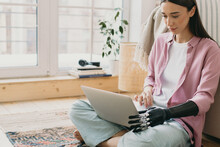 Close-up Picture Of Beautiful Charming Female In Pale Pink Silk Shirt Sitting On Floor On Colorful Carpet Holding Laptop On Knees With Prosthetic Bionic Hand Made Of Black Metal Mechanical Device