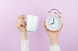 Human hand is holding coffee cup and pink alarm clock on pink background. It is coffee time in morning, concept with copy space.