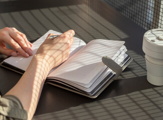 Poster - Female hands closeup taking notes in planner. Woman sitting at table with eco cup and writing plans or thoughts in office or cafe with sunshine. High quality photo