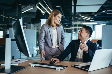 happy coworkers discuss project strategy by looking and pointing at laptop pc computer monitor screen. multiethnic business team in the office. confident mature asian man explaining young female