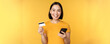 Online shopping. Cheerful asian girl holding credit card and smartphone, paying, order with mobile phone, standing over yellow background