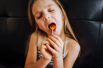 A beautiful, red-haired child with a sweet tooth sits on a black leather sofa with a lollipop in her hands and eats it.