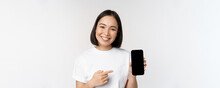 Smiling Asian Woman Pointing Finger At Smartphone Screen, Showing Application Interface, Mobile Phone Website, Standing Over White Background