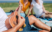 Unrecognizable Senior Man Playing Ukulele For His Family Sitting On A Blanket During An Excursion Outdoors