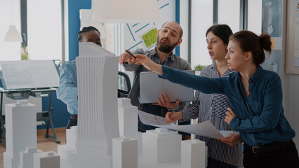 Group of colleagues meeting to analyze building model and maquette to plan blueprints. People working on architecture design with laptop, tablet and urban layout on table. Development