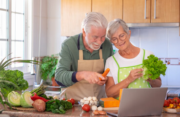 Attractive senior happy couple preparing vegetables together in the home kitchen searching menu at laptop computer. Caucasian elderly people enjoying healthy eating