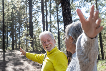 Attractive senior man looking at his wife walking in the forest holding her hand. Caucasian happy active elderly gray haired couple enjoying vacation freedom and nature hiking in footpath