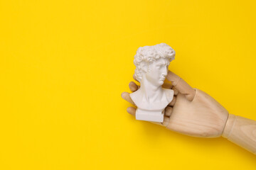 Wall Mural - Wooden hand holding David bust on yellow background. Minimal still life