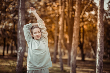 Attractive fit slim Caucasian woman with curly hair in sportswear is doing stretching exercise in the park or forest on a bright sunny day. Healthy lifestyle