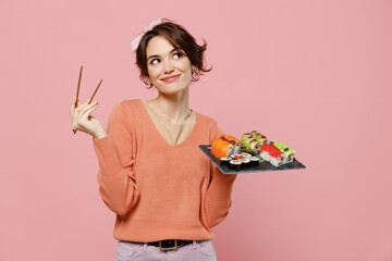 Wall Mural - Young minded pensive thoughtful fun woman in sweater hold in hand makizushi sushi roll served on black plate traditional japanese food look aside chopsticks isolated on plain pastel pink background