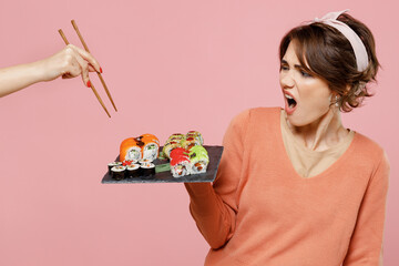 Wall Mural - Young disturbed woman in sweater hold in hand makizushi sushi roll served on black plate traditional japanese food do not want share give to another person isolated on plain pastel pink background