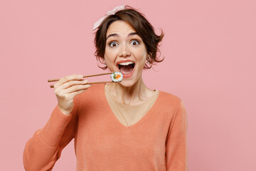 Wall Mural - Young excited happy cool fun caucasian woman 20s wear casual clothes hold in hand with chopsticks eat biting makizushi sushi roll traditional japanese food isolated on plain pastel pink background