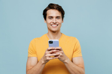 Young smiling happy cheerful man 20s wear yellow t-shirt hold in hand use mobile cell phone chatting browsing isolated on plain pastel light blue background studio portrait. People lifestyle concept