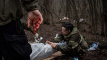 Hand Close-up With Blood On The Fingers And A Ring. Blurred Figures Of People In The Forest. First Aid In An Extreme Situation. Saving A Life In A Military Conflict. Tactical Medic, Victim, Rescue.