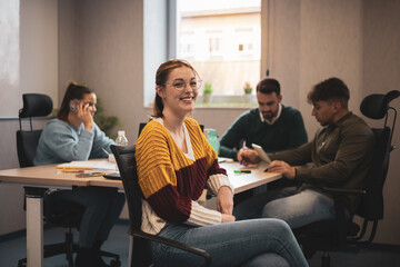 Portrait of a happy millennial business woman in a modern office. Businesswoman with glasses smiling and looking at the camera. Diverse and busy team working in the background. Coworking concept.