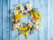 Easter Decoration. A Wreath Of Daffodils, Mimosa And Twigs With Colorful Quail Eggs On A Background Of Blue Wooden Boards. Copy Space