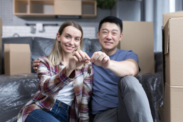 Portrait of a young couple man and woman looking at the camera and smiling holding the keys to a new apartment, multiracial family sitting on the bed of a new house hugging