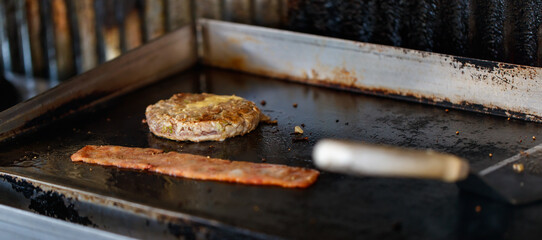 Closeup shot of delicious fresh baked burger bun with butter, minced cooked patty beef meat with margarine and pork bacon grilled on frying pan stove at dirty unsanitary local restaurant counter