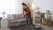 full length asian woman is slowly vacuuming the rug while doing housecleaning in a bright living room at home.