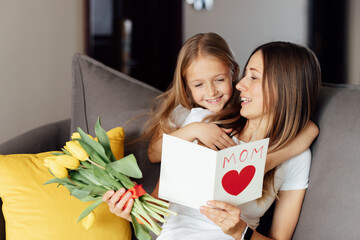 Child daughter congratulates mother and gives present card, gift and bouquet of flowers at home. Mom and girl smiling and hugging on couch. Happy mothers day celebration