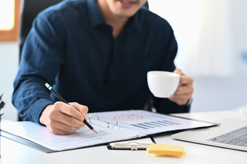 Wall Mural - Young businessman holding cup of coffee and checking financial reports at office desk.