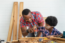 African Black Male Carpenter Man Father Teach Boy Kid Child Son To Be Carpenter Drilling Wood In Carpentry Workshop. Concept Hobby At Home.
