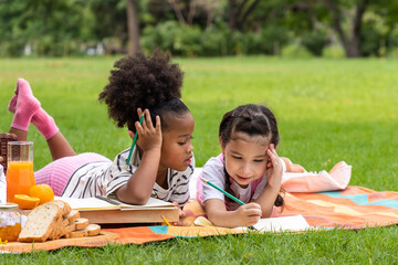 Two young girls multiracial lying and draw on lawn in the park on beautiful summer day, doing homework with friend.