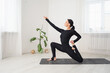 Woman in black sportswear practicing yoga doing anjaneyasana exercise with virasana, low lunge pose, sitting on a mat in a room by the window
