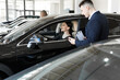 © Artem Zakharov - Side view of young beautiful woman sitting inside car and holding hand on steering wheel. She smiling and talking with manager of car dealership. Car agent representing inside of automobile.