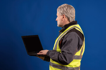 Poster - Experienced engineer. Gray-haired man with laptop. Man in yellow vest stands with laptop. Experienced engineer on blue background. Laptop with dark screen in hands of worker. Master with computer